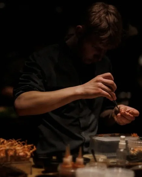 A young Alchemist chef plating with tweezers in the dim research kitchen